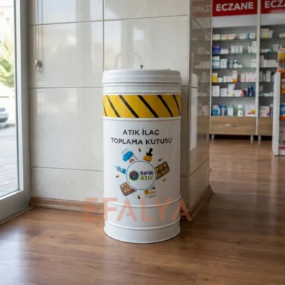 Modern bir eczane içerisinde, ilaç rafları yanında konumlandırılmış; üzerinde sarı-siyah ikaz şeridi, ilaç görselleri ve "ATIK İLAÇ TOPLAMA KUTUSU" yazısı bulunan beyaz silindirik metal kutu.