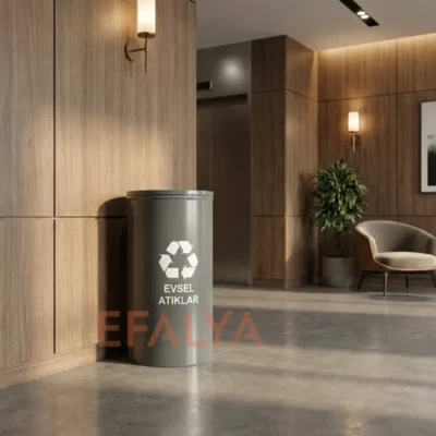Modern ve lüks bir otel holünde, ahşap panel duvar yanında konumlandırılmış; üzerinde beyaz geri dönüşüm sembolü ve "EVSEL ATIKLAR" yazısı bulunan silindirik gri metal atık kutusu.