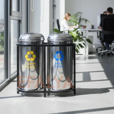 Gün ışığı alan modern bir ofis ortamında, pencere önünde konumlandırılmış; plastik (sarı) ve kağıt (mavi) ayrıştırma bölmelerine sahip, siyah metal şaseli 2'li paslanmaz çelik geri dönüşüm ünitesi.