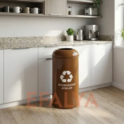Modern bir ofis mutfağında, beyaz dolaplar önünde konumlandırılmış; üzerinde beyaz geri dönüşüm sembolü ve "BİYOBOZUNUR ATIKLAR" yazısı bulunan silindirik kahverengi metal atık kutusu.