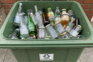 Yeşil renkli cam atık geri dönüşüm kutusu içerisinde biriktirilmiş şeffaf, yeşil ve kahverengi cam şişeler, soda şişeleri ve cam kavanozlar; üzerinde "Cam Atıklar" yazan toplama ünitesi.