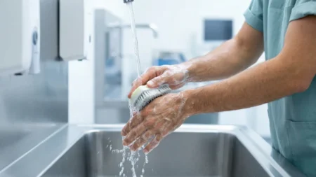 Ameliyathane ortamında paslanmaz çelik cerrahi evyede antiseptik sünger kullanarak ameliyat öncesi cerrahi el yıkama protokolünü uygulayan cerrah.